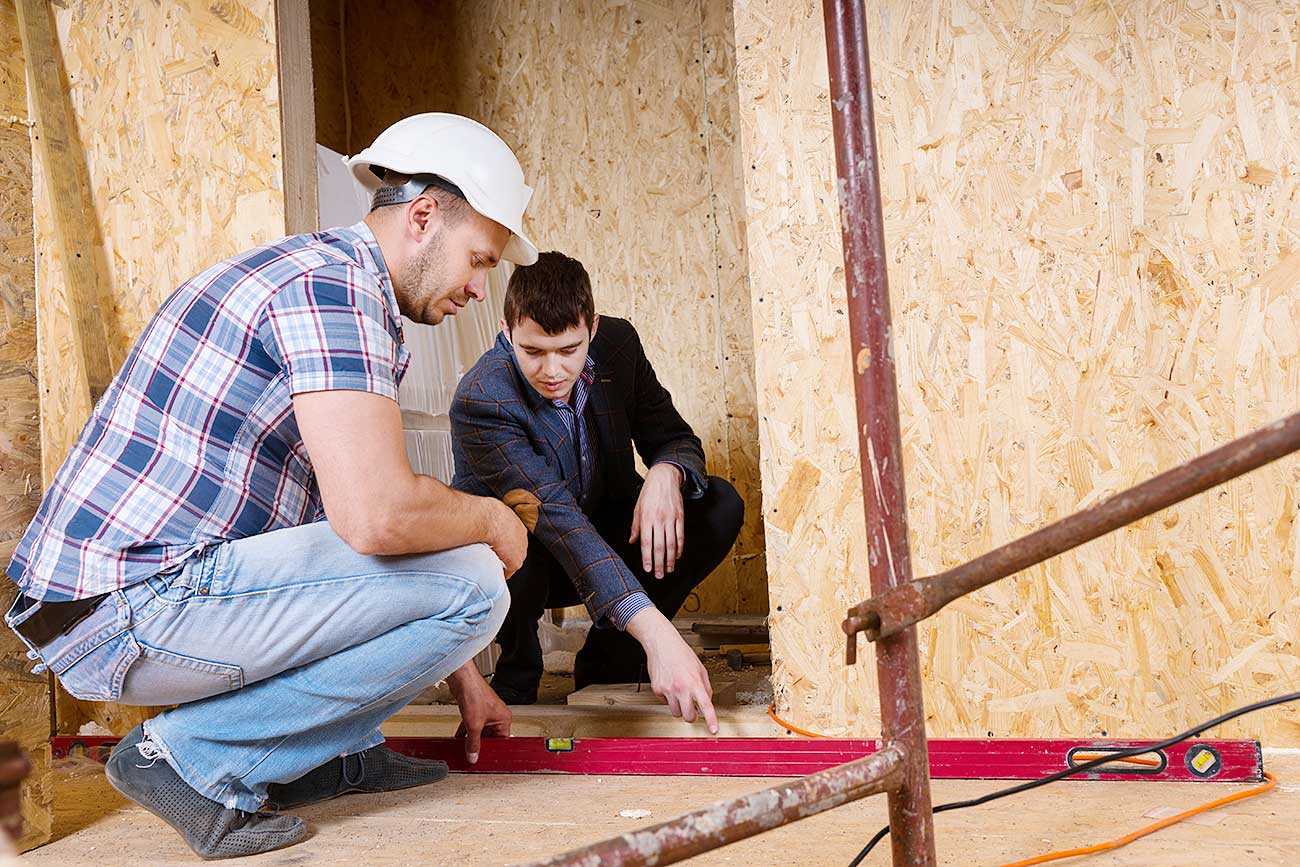 A construction worker and a surveyor discuss options during a build. They are crouched down looking at a spirit level. The background is the interior of a building under construction.